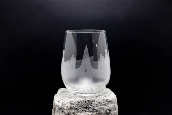 A clear glass with an etched city skyline design on it, placed on a rock against a dark background.