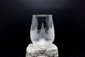 A clear glass with an etched city skyline design on it, placed on a rock against a dark background.