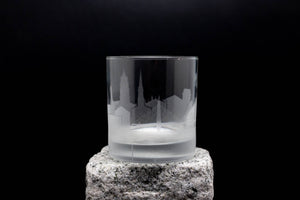 A frosted glass with an etched city skyline design sits on a rock against a black background.