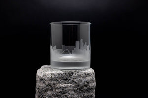 A clear glass with an etched city skyline design sits on a rock against a dark background.