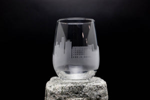 A glass with an etched city skyline design on it, placed on a rock.