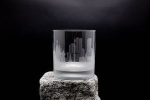 A frosted glass with an etched city skyline design sits on a rock against a dark background.